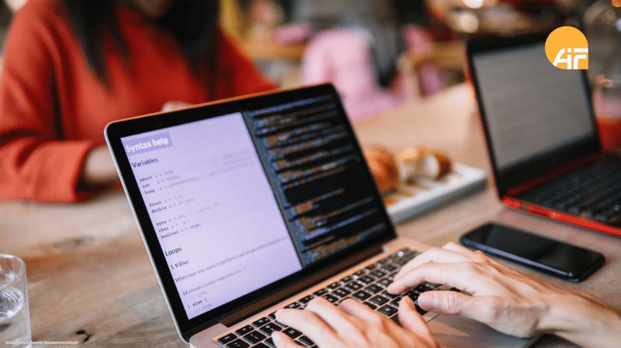 Ein offener Laptop, an dem eine Person arbeitet. Im Hintergrund sieht man andere arbeitende Personen.