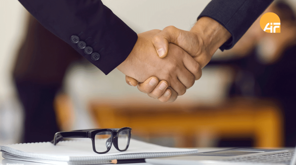 Zwei Personen, die sich die Hände schütteln. Im Vordergrund liegen mehrere Blöcke und eine Brille.